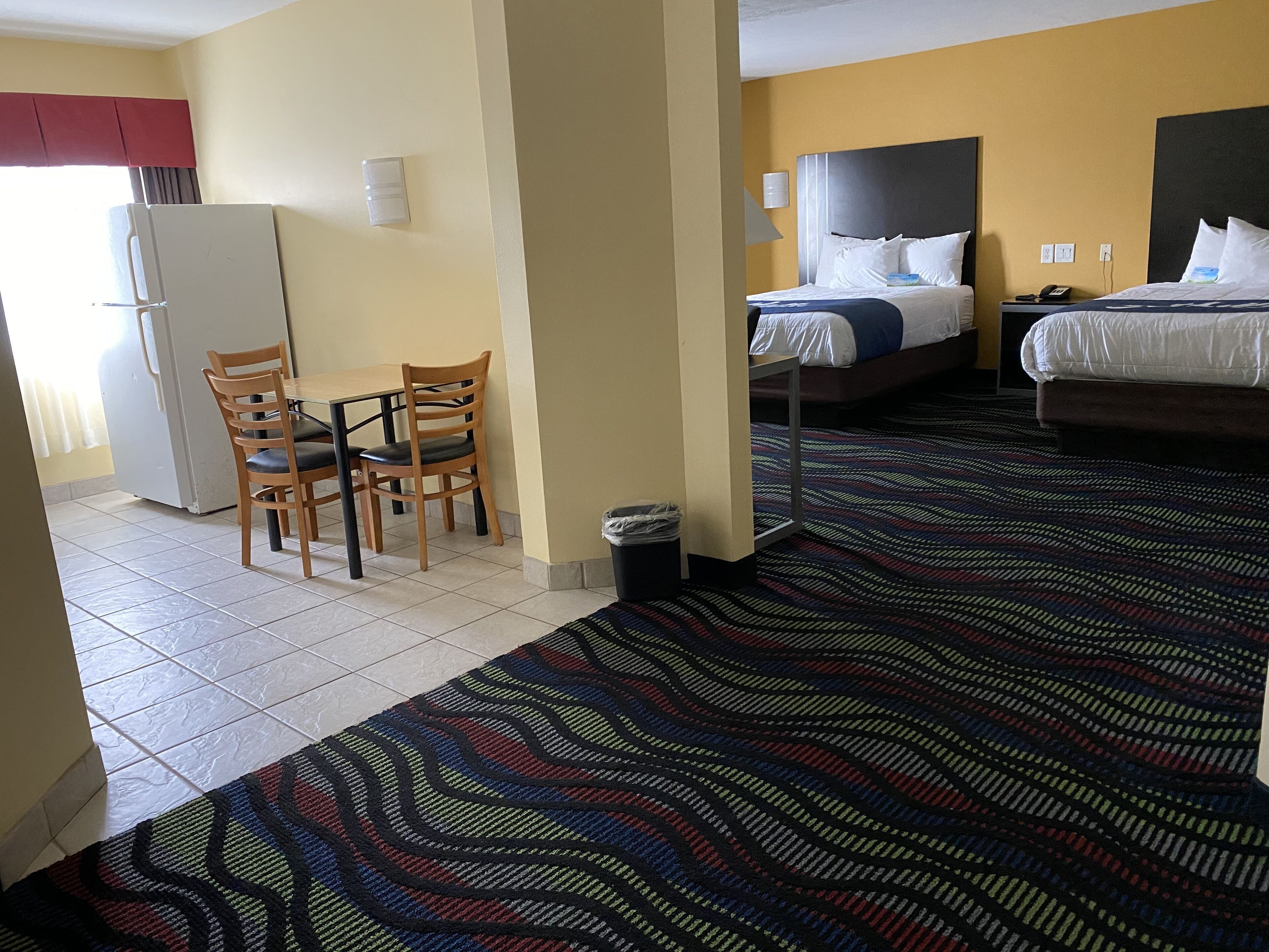 A hotel room with a bed, a desk with chairs, a refrigerator, and a patterned carpet.