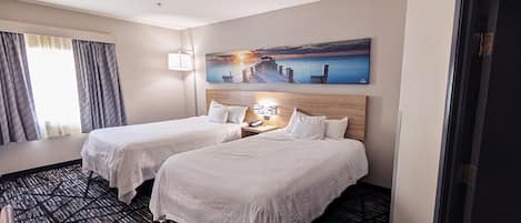 A hotel room with two beds, a painting of a sunset pier, a desk with a lamp, and a window with curtains.