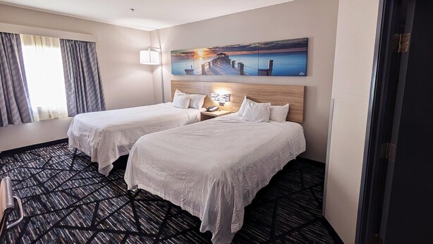A hotel room with two beds, a large window with curtains, a painting above the beds, and a carpeted floor. - Days Inn & Suites by Wyndham Augusta Near Fort Eisenhower (Grovetown)