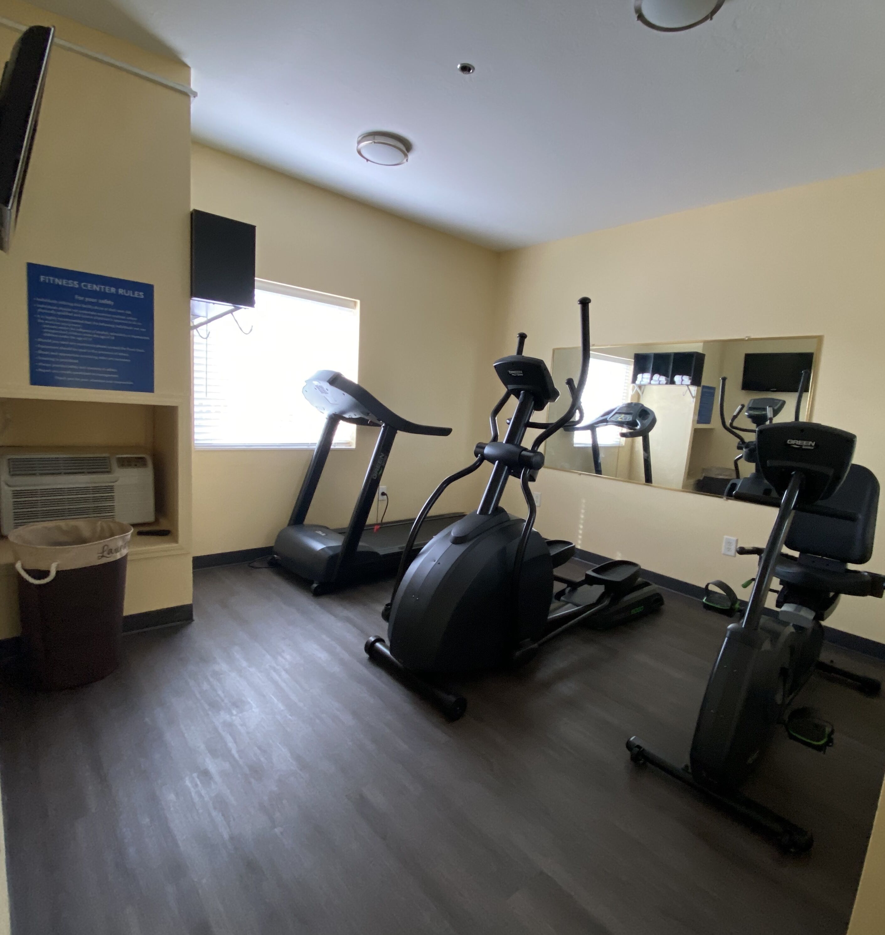 Centro de fitness com esteiras, elíptica e bicicleta ergométrica. Há um cesto de lixo e uma placa com regras.