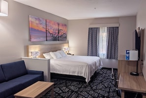 A hotel room with a bed, a sofa, a desk, a lamp, a TV, and a painting of a sunset. - Days Inn & Suites by Wyndham Augusta Near Fort Eisenhower (Grovetown)