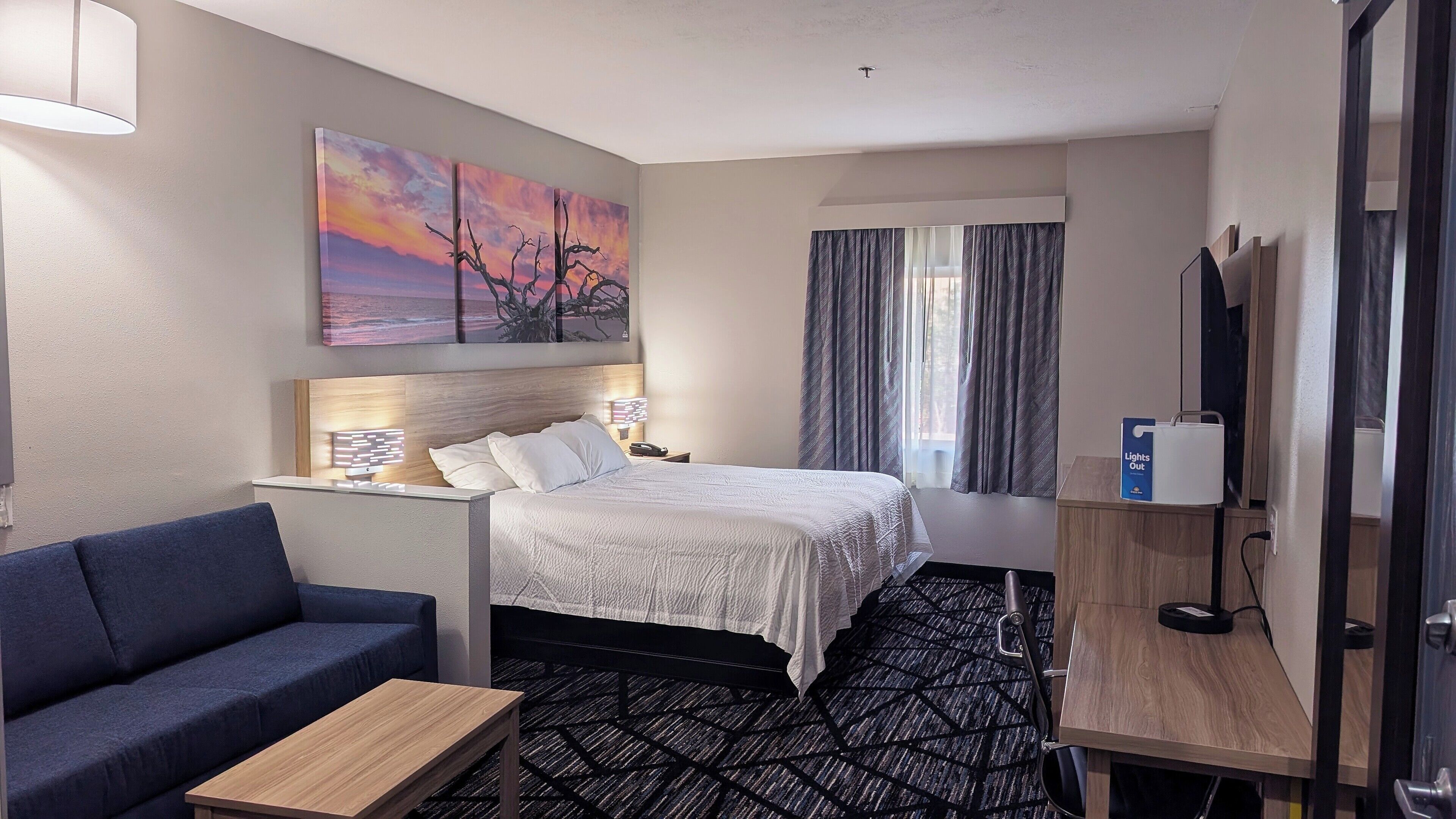 Una habitación de hotel con cama, sofá azul, mesita, mesita de noche con lámpara, un cuadro en la pared y ventana con cortinas.