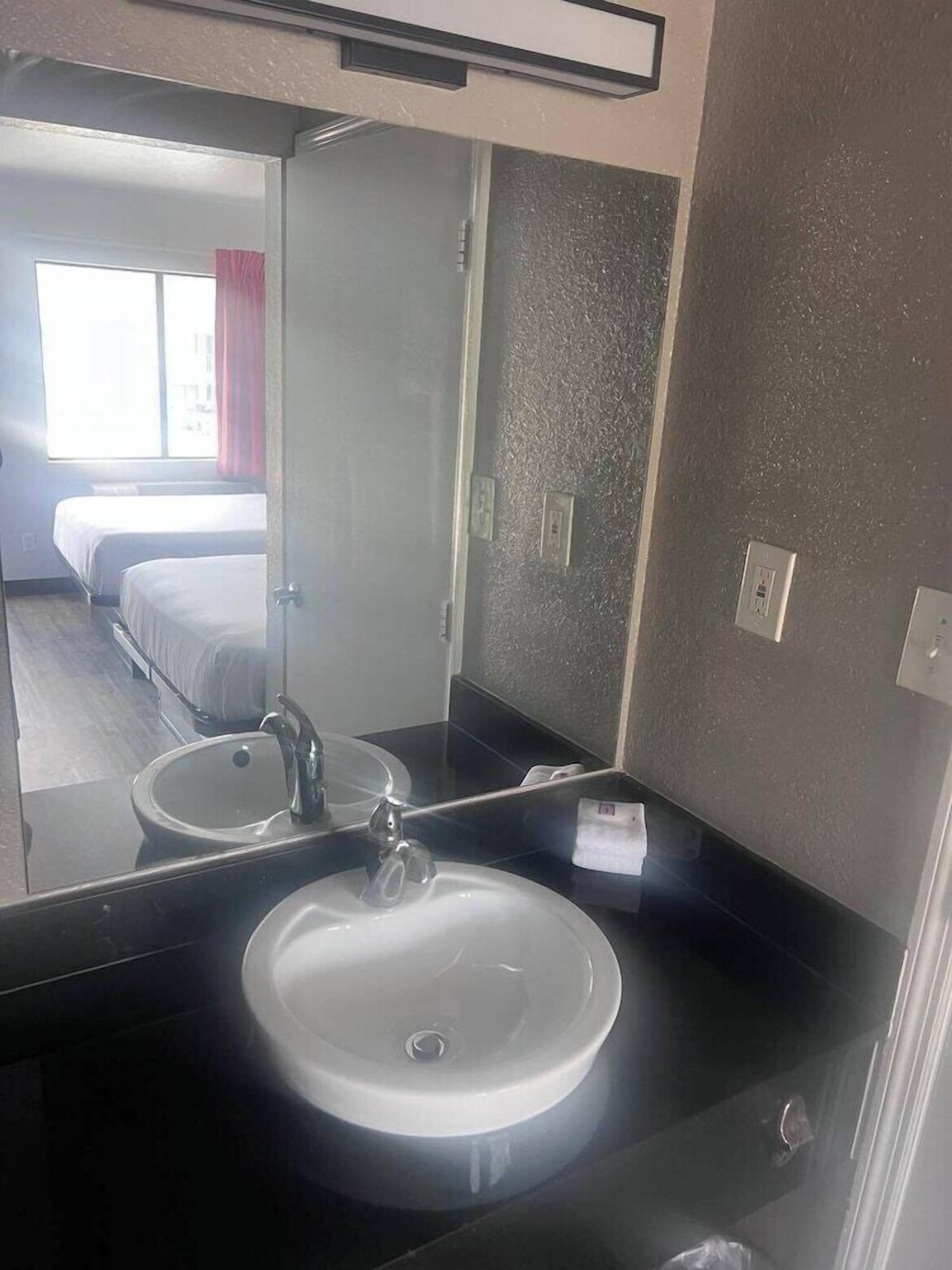 A hotel bathroom with a large mirror, two sinks, and a bed visible in the background.