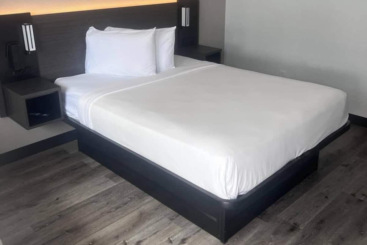 A modern hotel room with a large bed, wooden flooring, and built-in nightstands with bedside lamps.