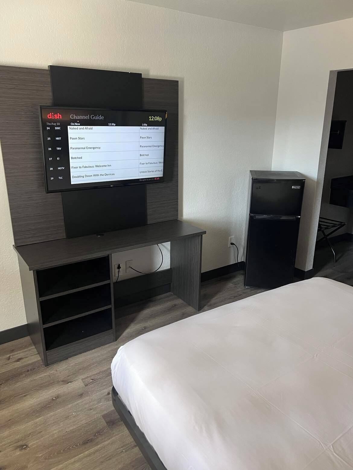 A hotel room with a flat-screen TV mounted on the wall displaying a Channel Guide. There is a bed with white bedding, a small refrigerator, and a wooden entertainment unit with shelves.