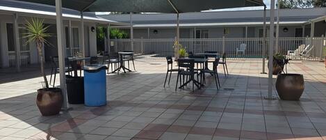 Un espacio de descanso al aire libre con mesas y sillas, plantas en macetas y una piscina al fondo.