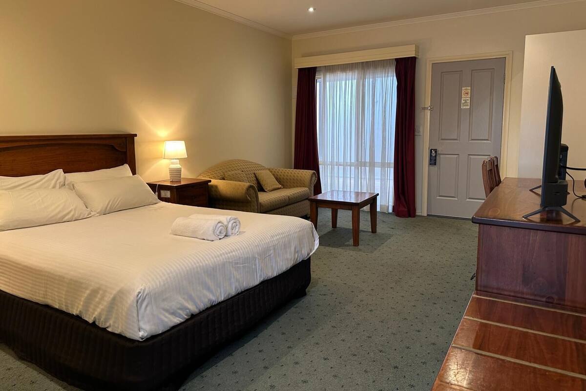 Ein Hotelzimmer mit Bett, Sessel, Tisch, Fernseher und einem Fenster mit Vorhängen.