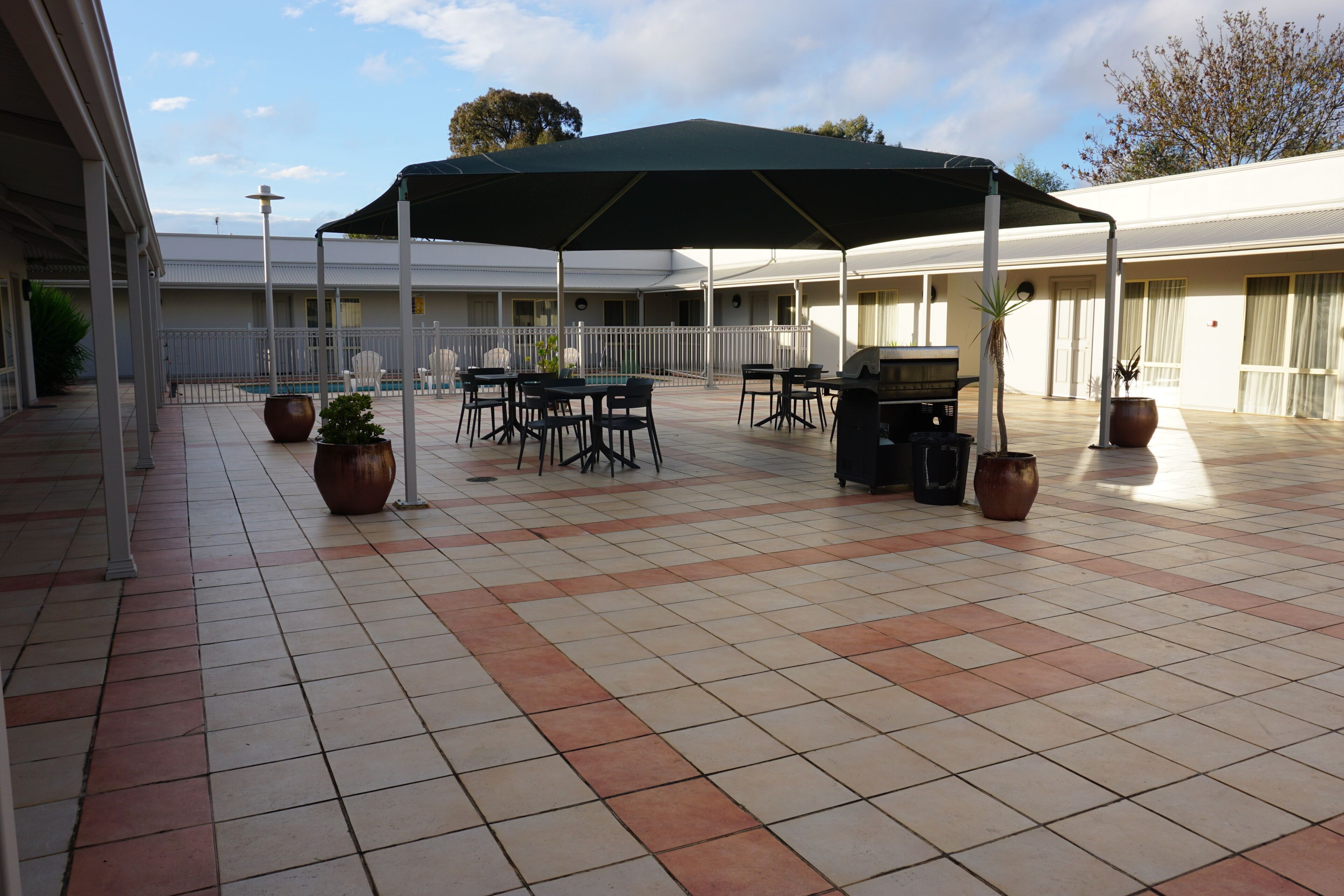 Une terrasse extérieure couverte, dotée d’un sol carrelé, de tables, de chaises, d’un barbecue et de plantes en pot.