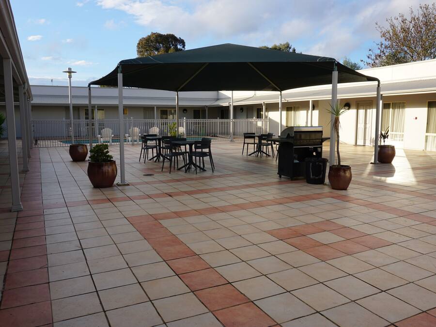 Une terrasse extérieure couverte, dotée d’un sol carrelé, de tables, de chaises, d’un barbecue et de plantes en pot.
