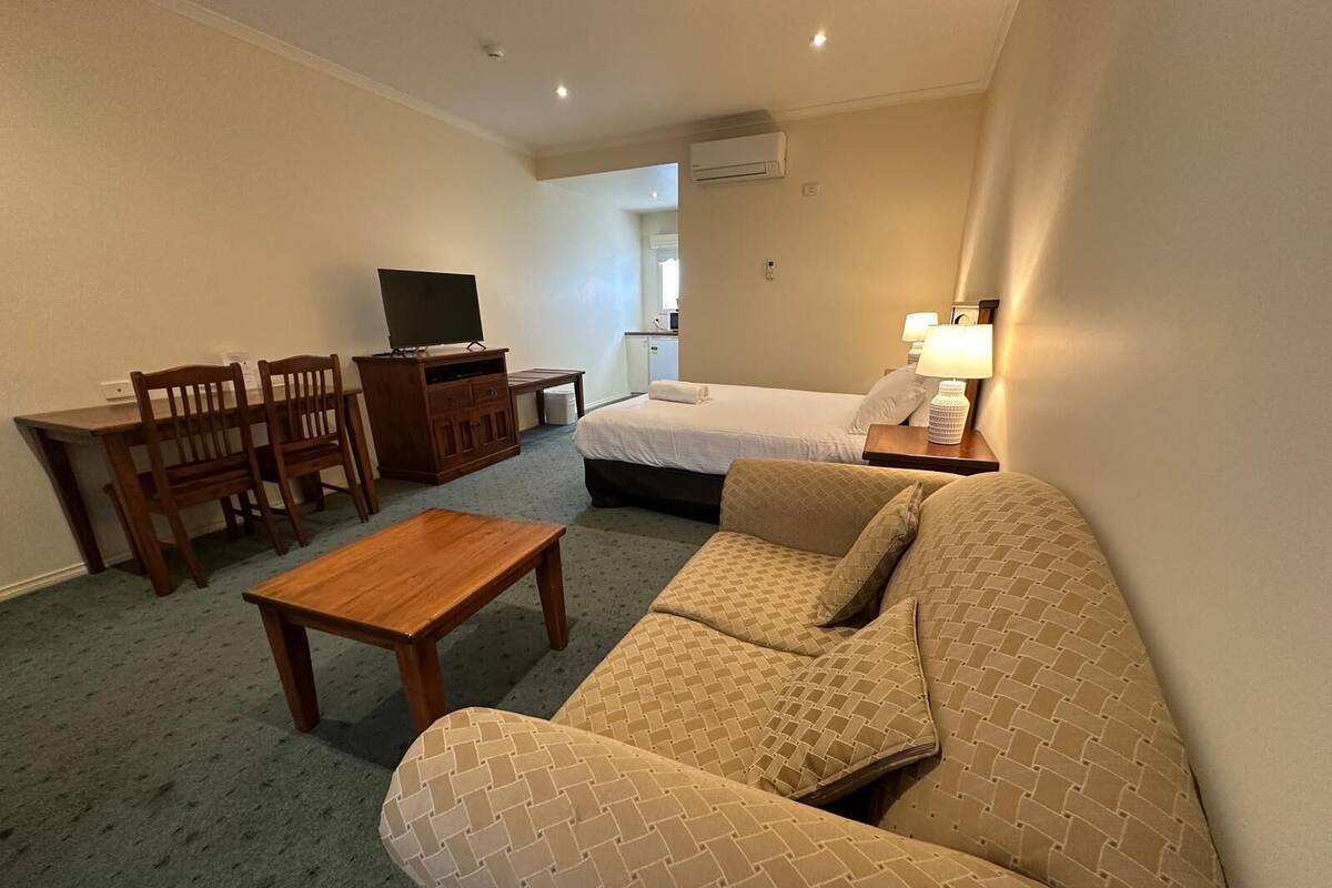 Ein Hotelzimmer mit einer Couch, einem kleinen Holztisch, einem Fernseher, einem Bett und einem Schreibtisch mit Stühlen.