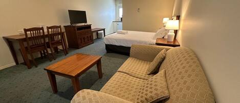 Une chambre d’hôtel avec un canapé, une petite table en bois, une télévision, un lit et un bureau avec des chaises.