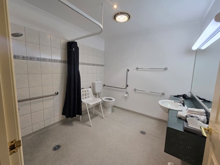 Un baño con paredes de azulejos blancos, inodoro, lavamanos, zona de ducha y barras de apoyo.