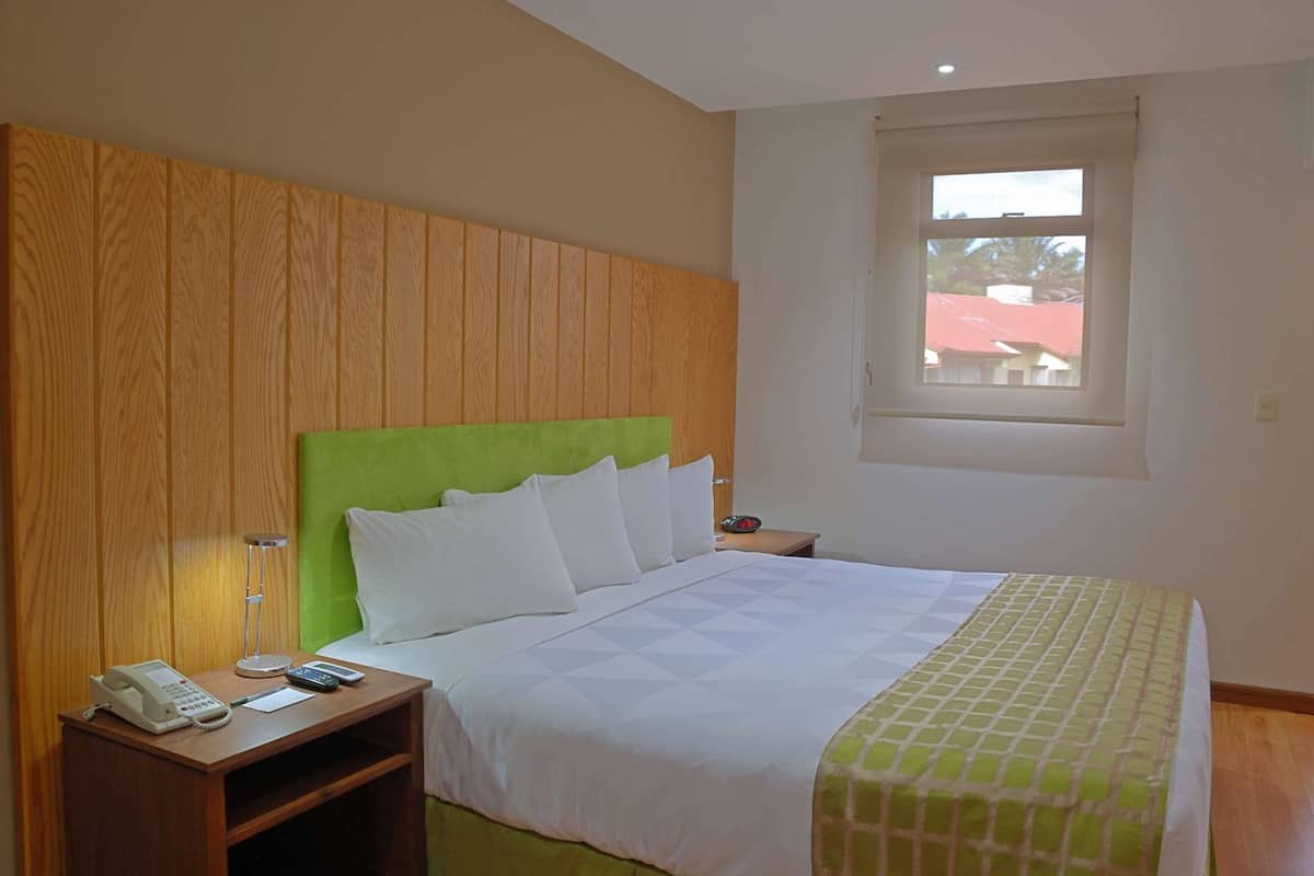 Ein Hotelzimmer mit Holz-Kopfteil, einem Bett mit weißer Bettwäsche, einem Nachttisch mit Telefon und Lampe sowie einem Fenster mit Blick auf Gebäude.