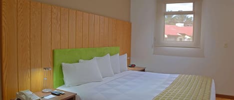 A hotel room with a wooden headboard, a bed with white bedding, a bedside table with a phone and lamp, and a window with a view of buildings.