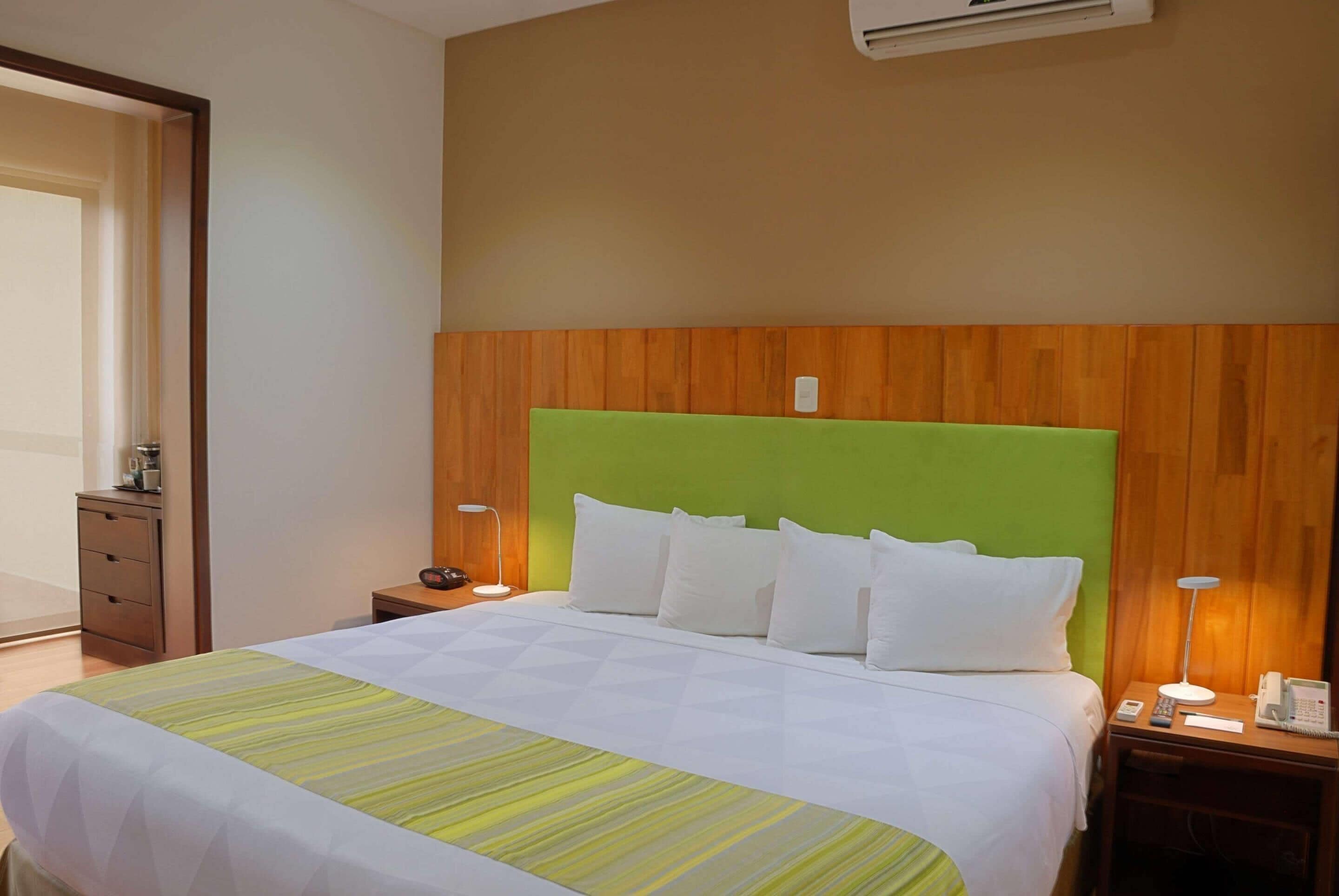 A hotel room with a bed, bedside tables, a nightstand, an air conditioning unit, and a wooden headboard.