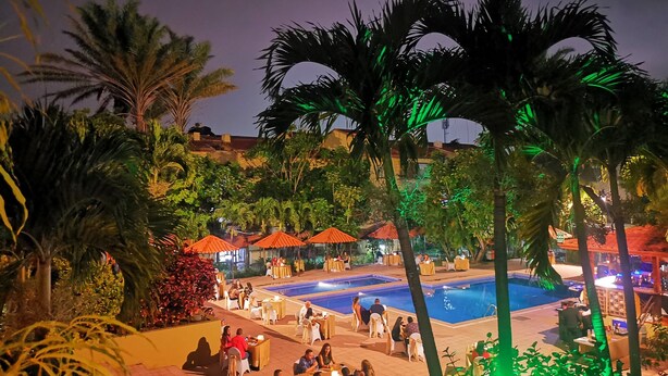A nighttime outdoor pool area with multiple dining tables and umbrellas, surrounded by palm trees and a pool.