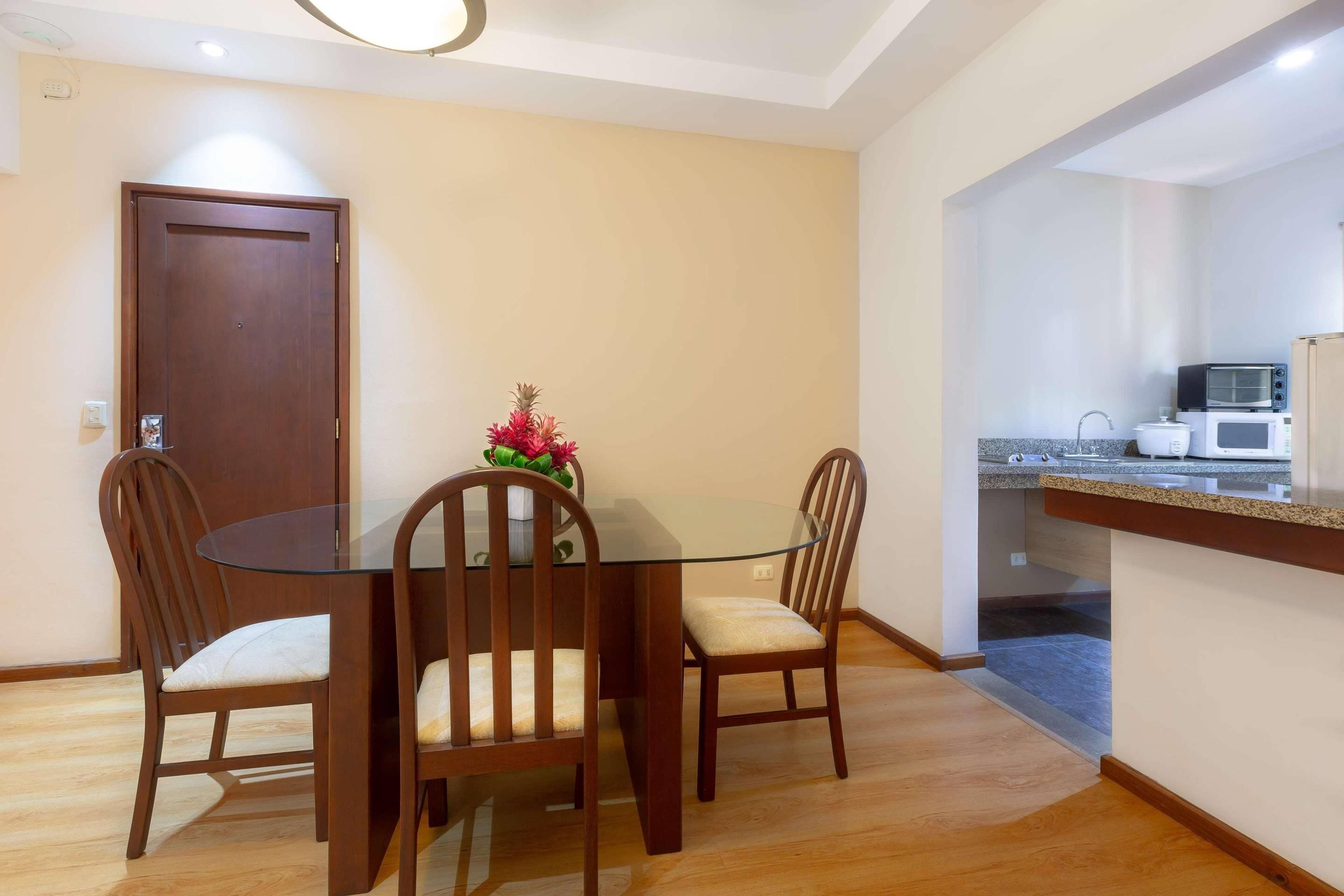 A dining area with a glass-top table and wooden chairs, a kitchen with a microwave and toaster, and a door in the background.