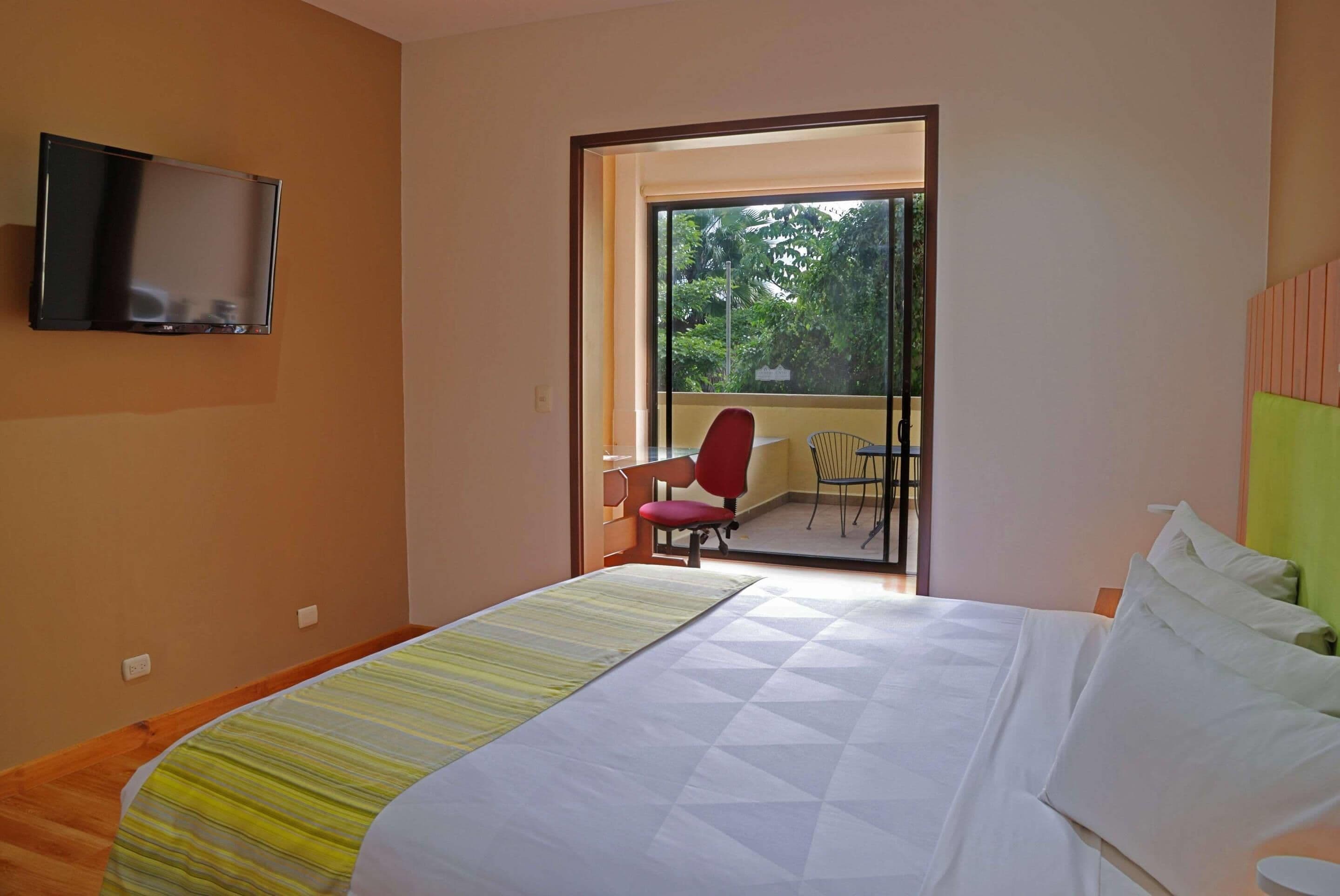 A hotel room with a bed, a TV mounted on the wall, a red chair, and a balcony with a table and chair.
