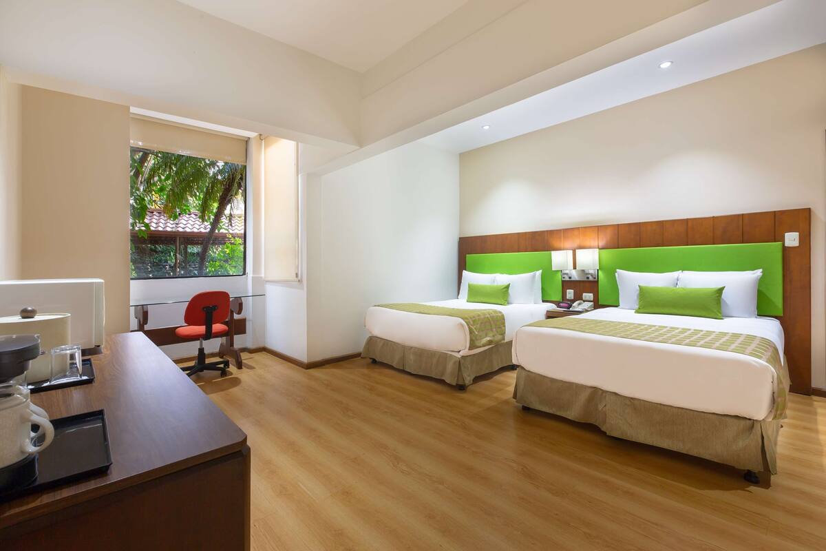 Ein Hotelzimmer mit zwei Betten, einem Schreibtisch, einem Sessel und Blick auf Grünflächen.