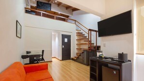 A modern living room with a wooden staircase, a flat-screen TV mounted on the wall, and a compact kitchen area with a refrigerator and a coffee machine.