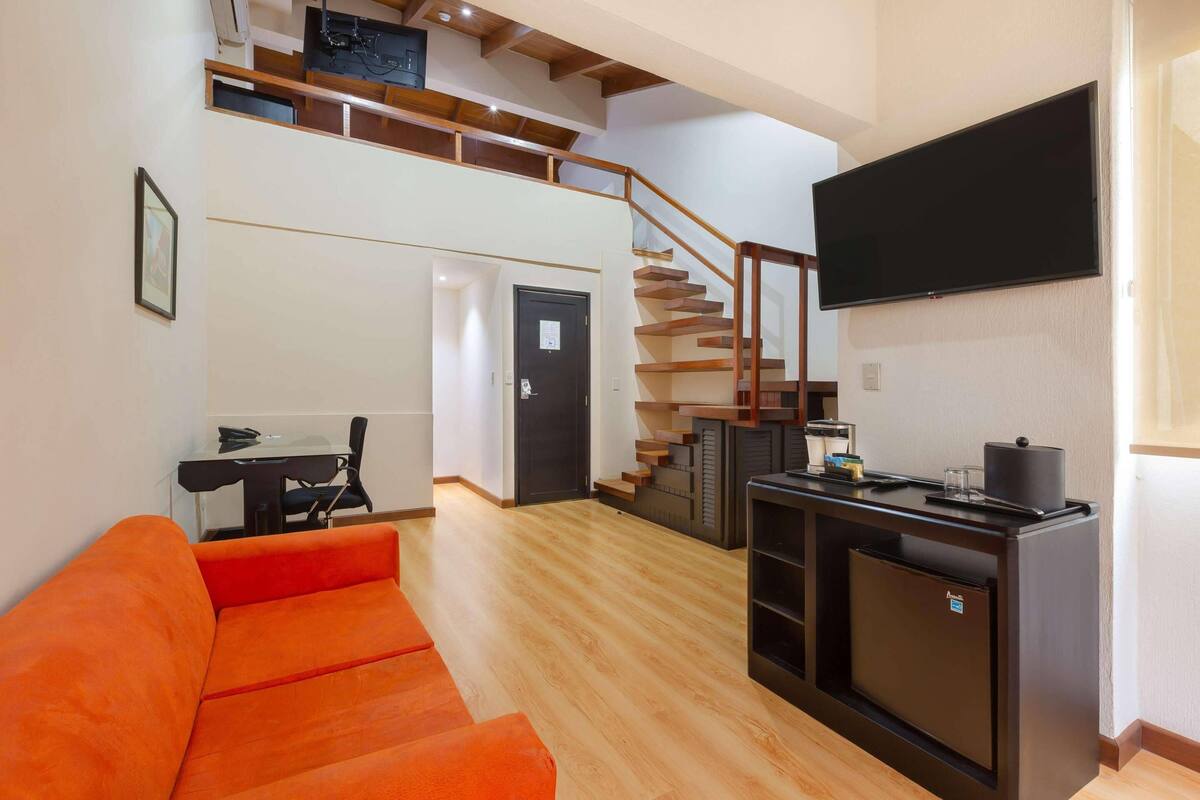 Ein modernes Wohnzimmer mit einer Holztreppe, einem Wand-Fernseher und einer kleinen Küchenzeile mit Kühlschrank und Kaffeemaschine.