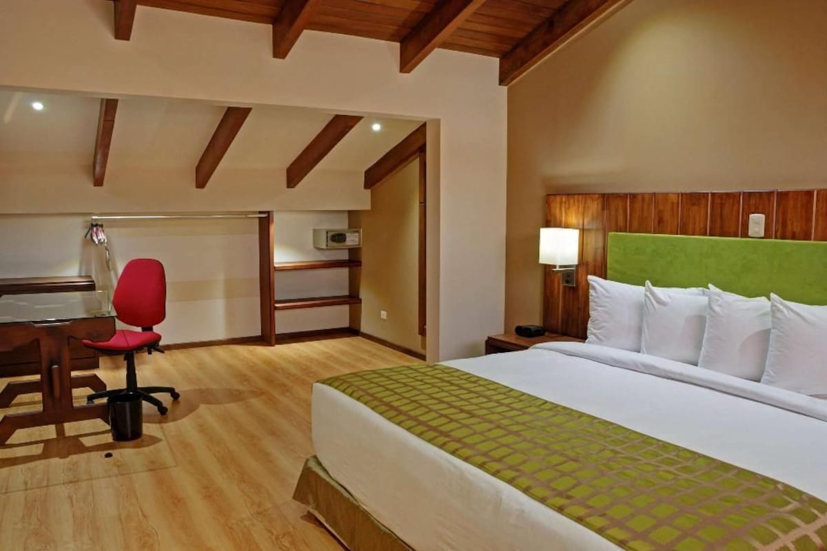 A hotel room with a bed, a desk, a red chair, a nightstand, and a green headboard.