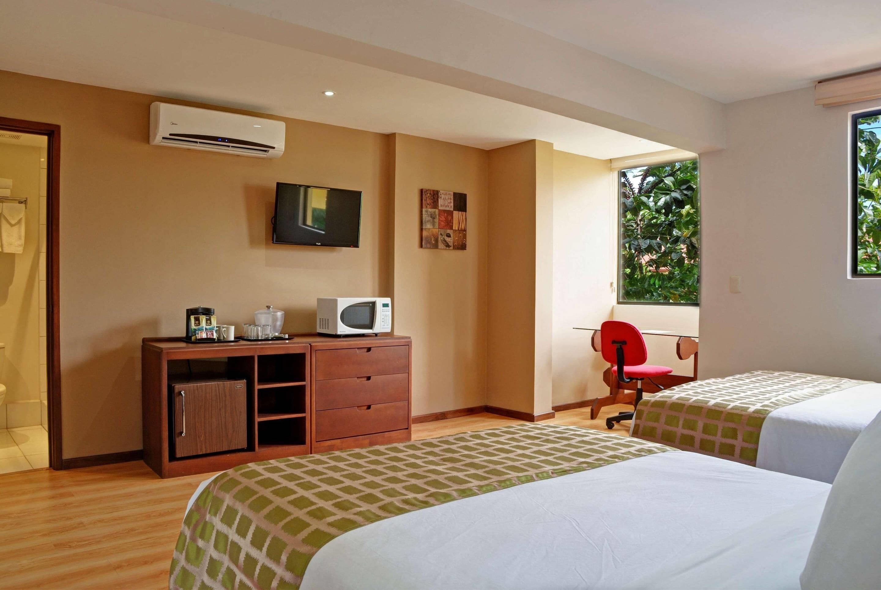 Habitación de hotel con dos camas, televisión, microondas, un mueble pequeño y ventana con vista a zonas verdes.