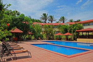 A swimming pool with lounge chairs and umbrellas, surrounded by palm trees and a building with a red roof.
