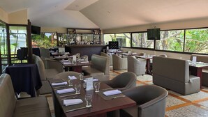 A restaurant with multiple dining areas, a bar, and large windows offering a view of greenery.
