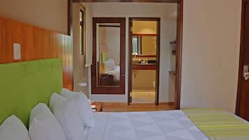 Quarto de hotel com uma cama, cabeceira verde, banheiro visível por uma porta aberta e piso de madeira.