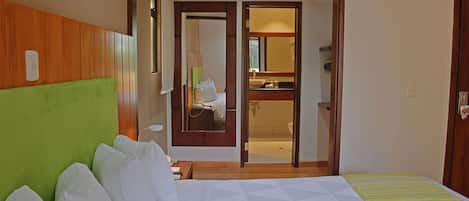 A hotel room with a bed, a green headboard, a bathroom visible through an open door, and a wooden floor.