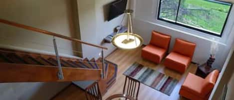 A modern living room with a staircase, orange sofas, a round table, and a large window with a view of greenery.