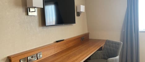 A hotel room with a wooden desk, a flat-screen TV mounted on the wall, a chair, and a window with curtains.
