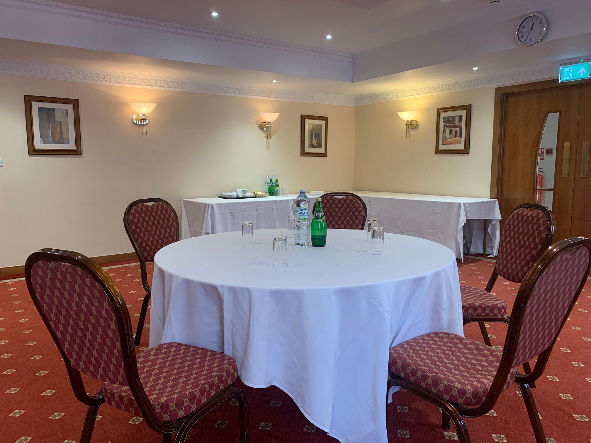 A conference room with a round table, four chairs, and a white tablecloth. There are wall-mounted light fixtures, framed pictures, and a clock on the wall.