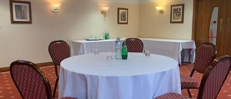 A conference room with a round table, four chairs, and a white tablecloth. There are wall-mounted light fixtures, framed pictures, and a clock on the wall.