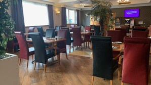 Een restaurant met houten vloeren, rode en zwarte stoelen en een televisie waarop 'Smooth Radio' te zien is.