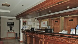 Een receptiebalie met een houten afwerking, een bord met de naam Ramada, en pijlen die naar faciliteiten wijzen zoals de congreszaal en het restaurant.