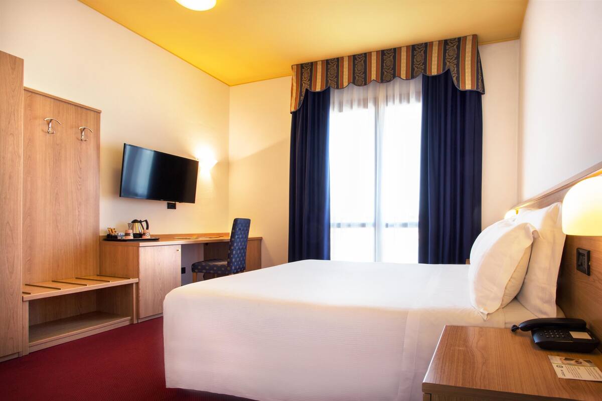 Habitación de hotel con una cama grande, un escritorio con silla, televisor y ventana con cortinas.