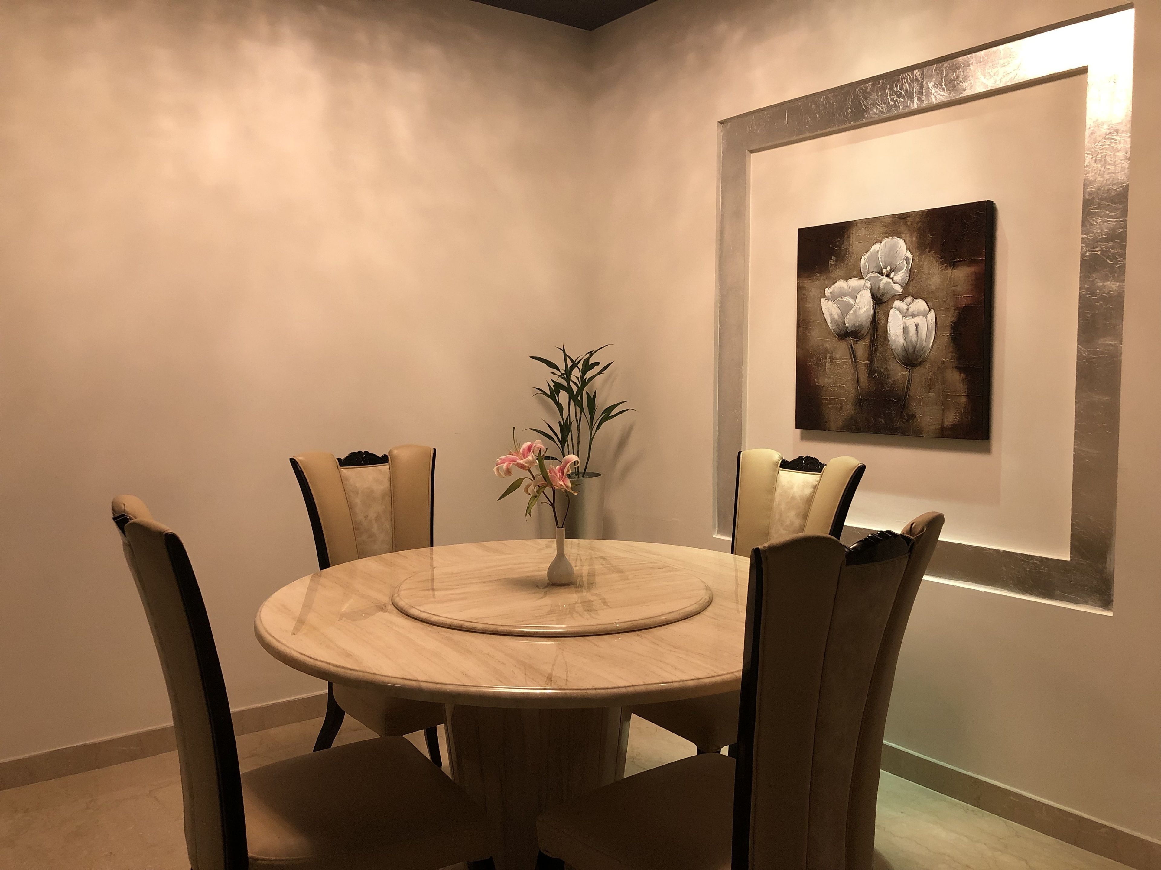 A dining area with a round table, four chairs, a vase with flowers, and a framed artwork on the wall.