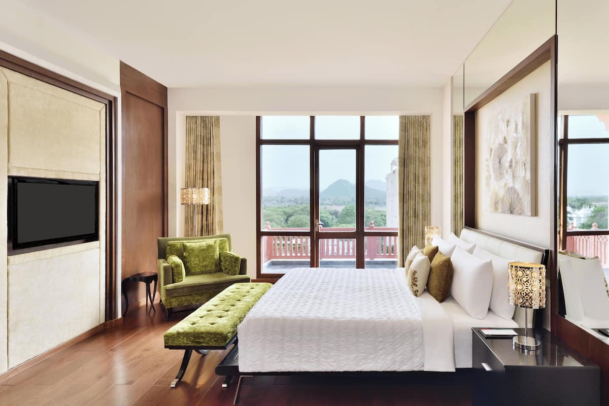 Quarto de hotel com uma cama grande, uma poltrona verde, uma mesinha, televisão e varanda com vista para a cidade.