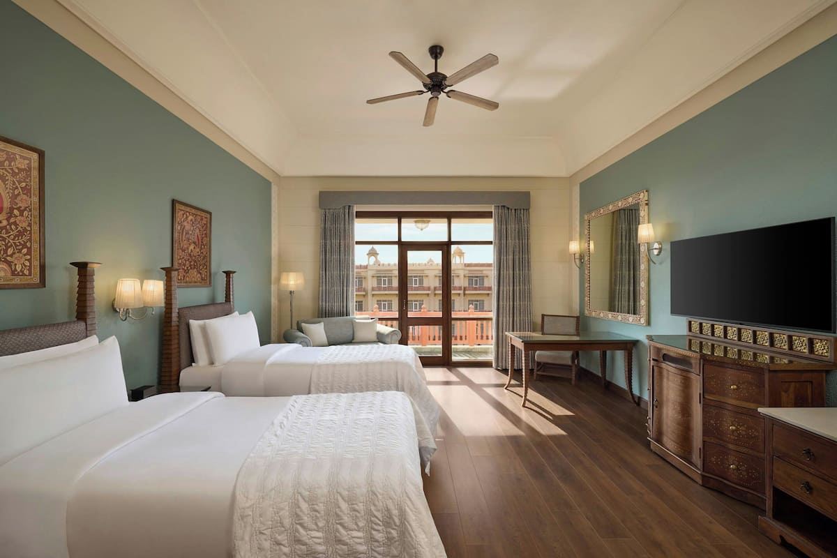 Quarto de hotel com duas camas, uma escrivaninha, televisão e varanda com vista para edifícios.