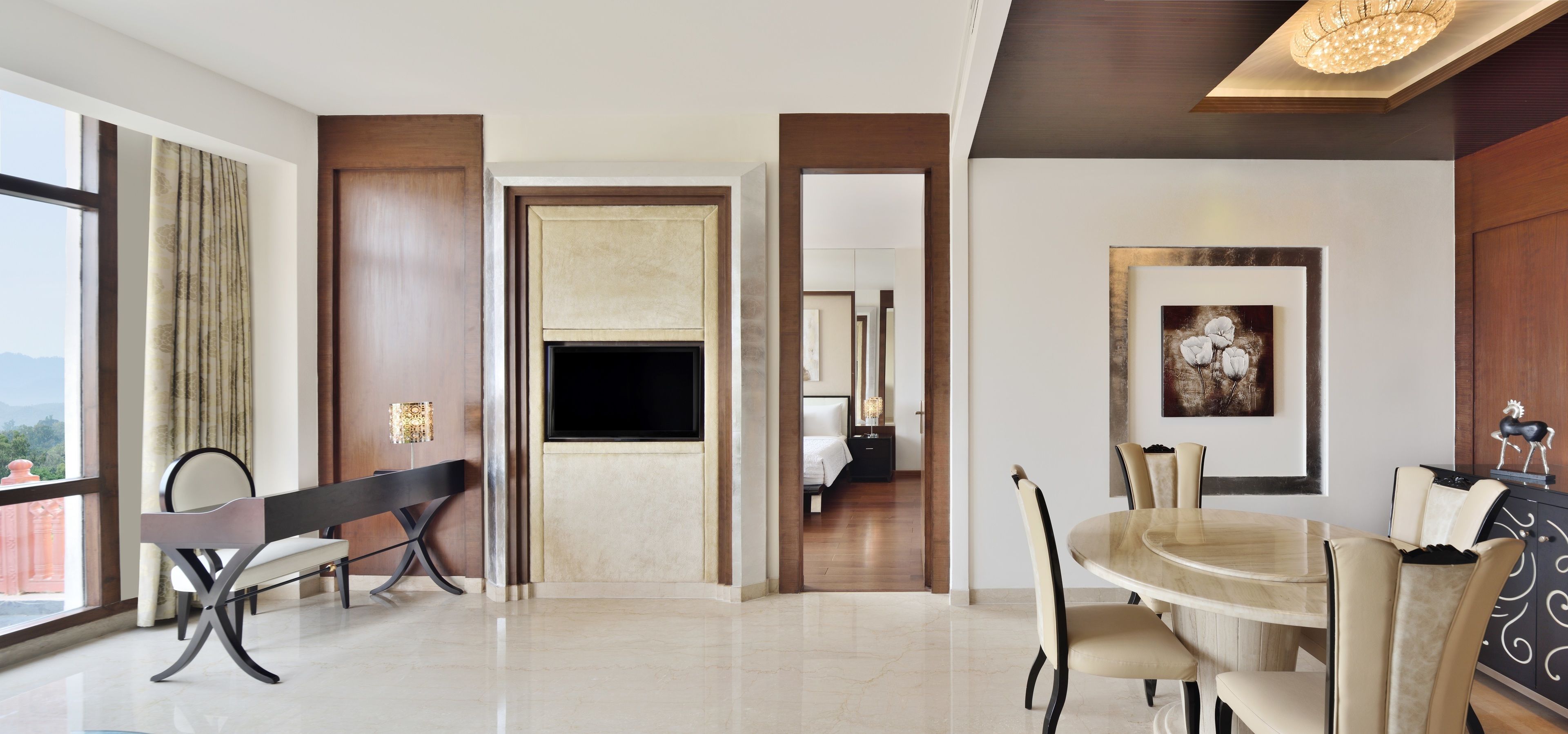 A modern hotel room with a large window, a flat-screen TV, a dining area with a round table and chairs, and a bedroom visible through an open door.