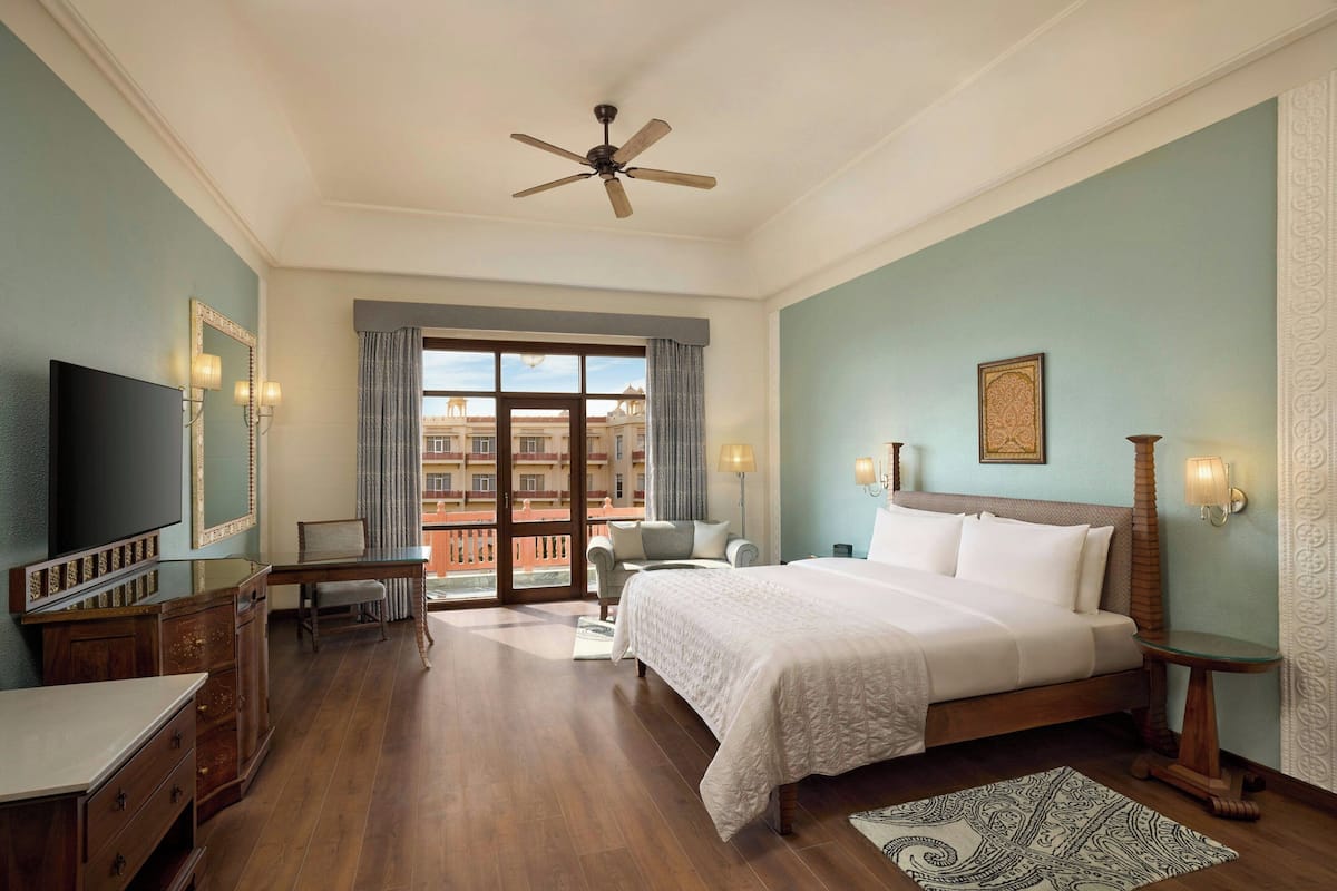 Quarto de hotel com cama, escrivaninha, cadeira, televisão e varanda com vista para edifícios.