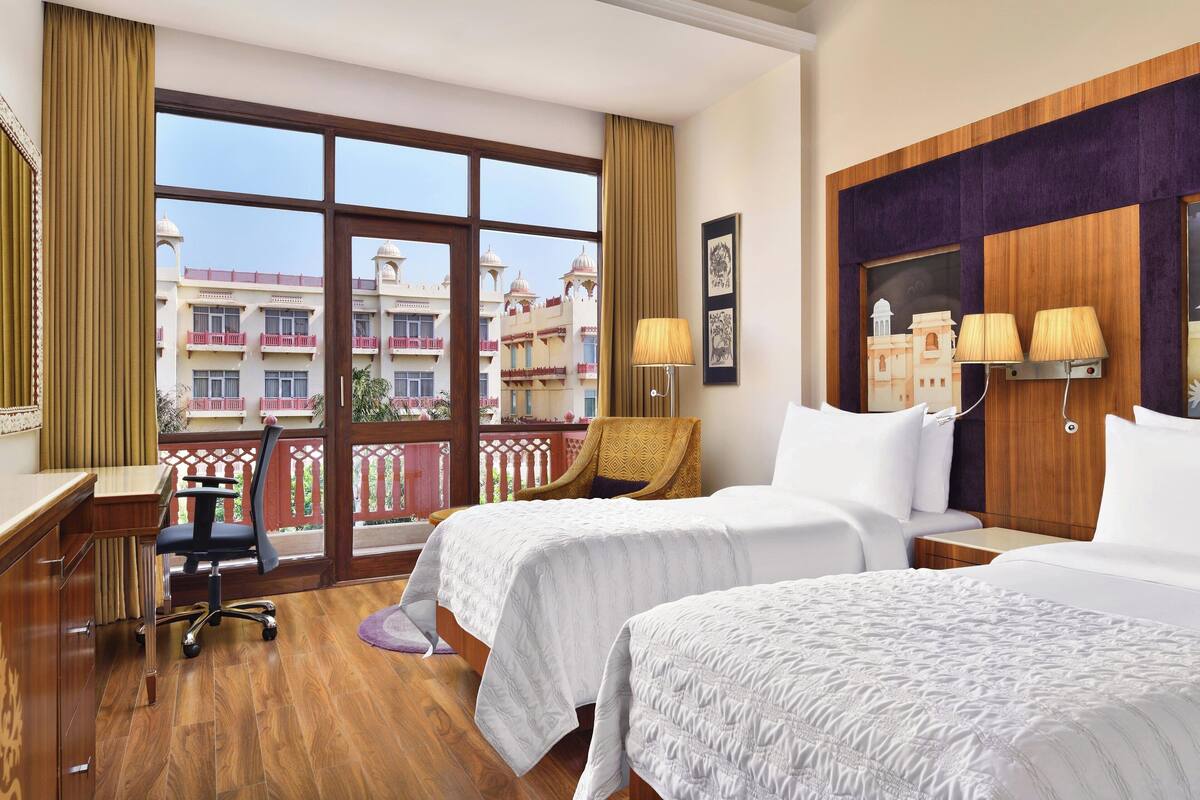 Quarto de hotel com duas camas, uma escrivaninha e uma cadeira. Há uma janela ampla com vista para um edifício com cúpulas.