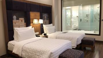 Quarto de hotel com duas camas, uma mesa de cabeceira com abajur, um espelho grande e vista para o banheiro.