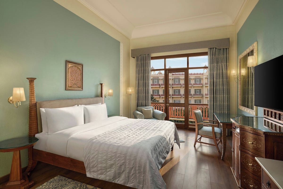 Quarto de hotel com uma cama grande, uma escrivaninha, uma cadeira, uma televisão e varanda com vista para edifícios.