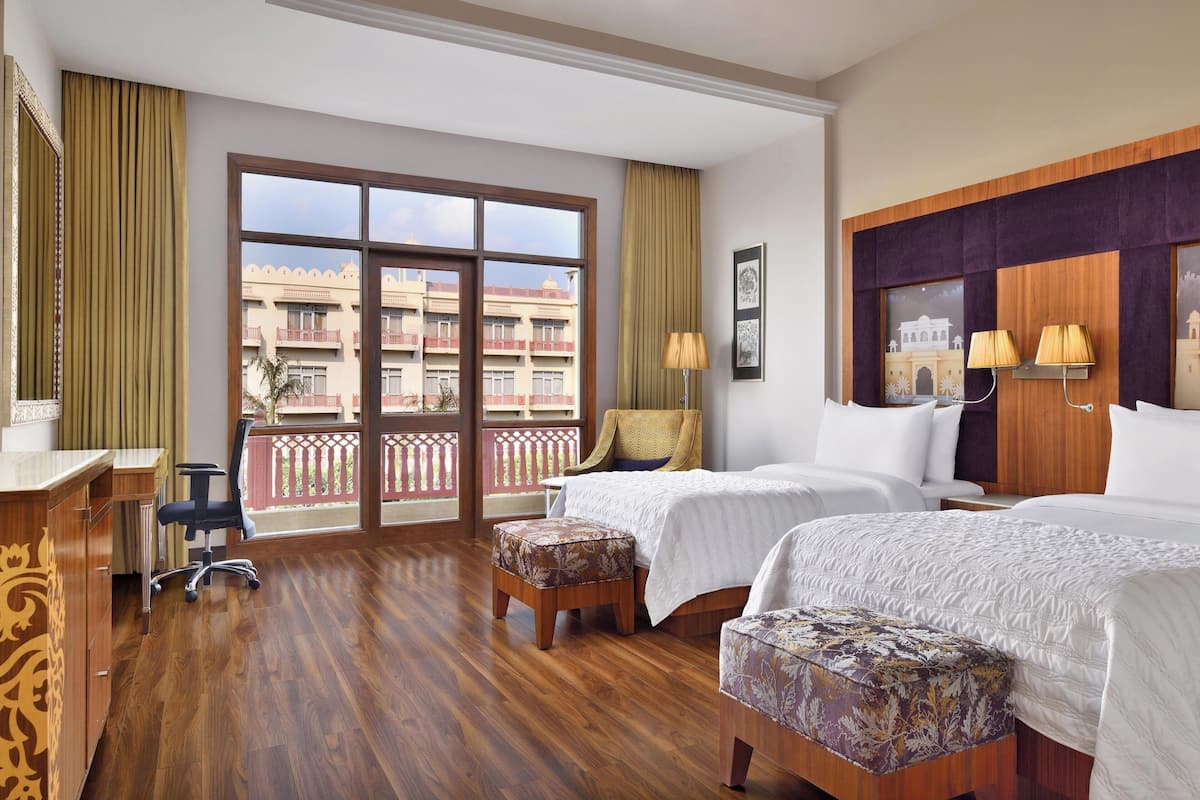 Quarto de hotel com duas camas, uma mesa, uma cadeira e varanda com vista para edifícios.