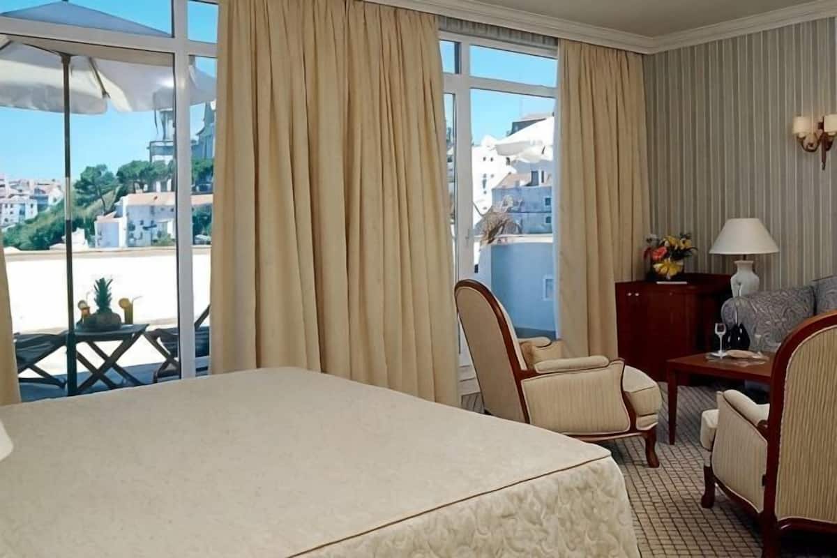 Habitación de hotel con cama, dos sillas, un escritorio con lámpara y vista a la ciudad a través de amplias ventanas.