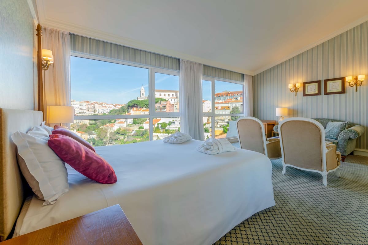 A hotel room with a large bed, two chairs, a desk, and a view of a cityscape.