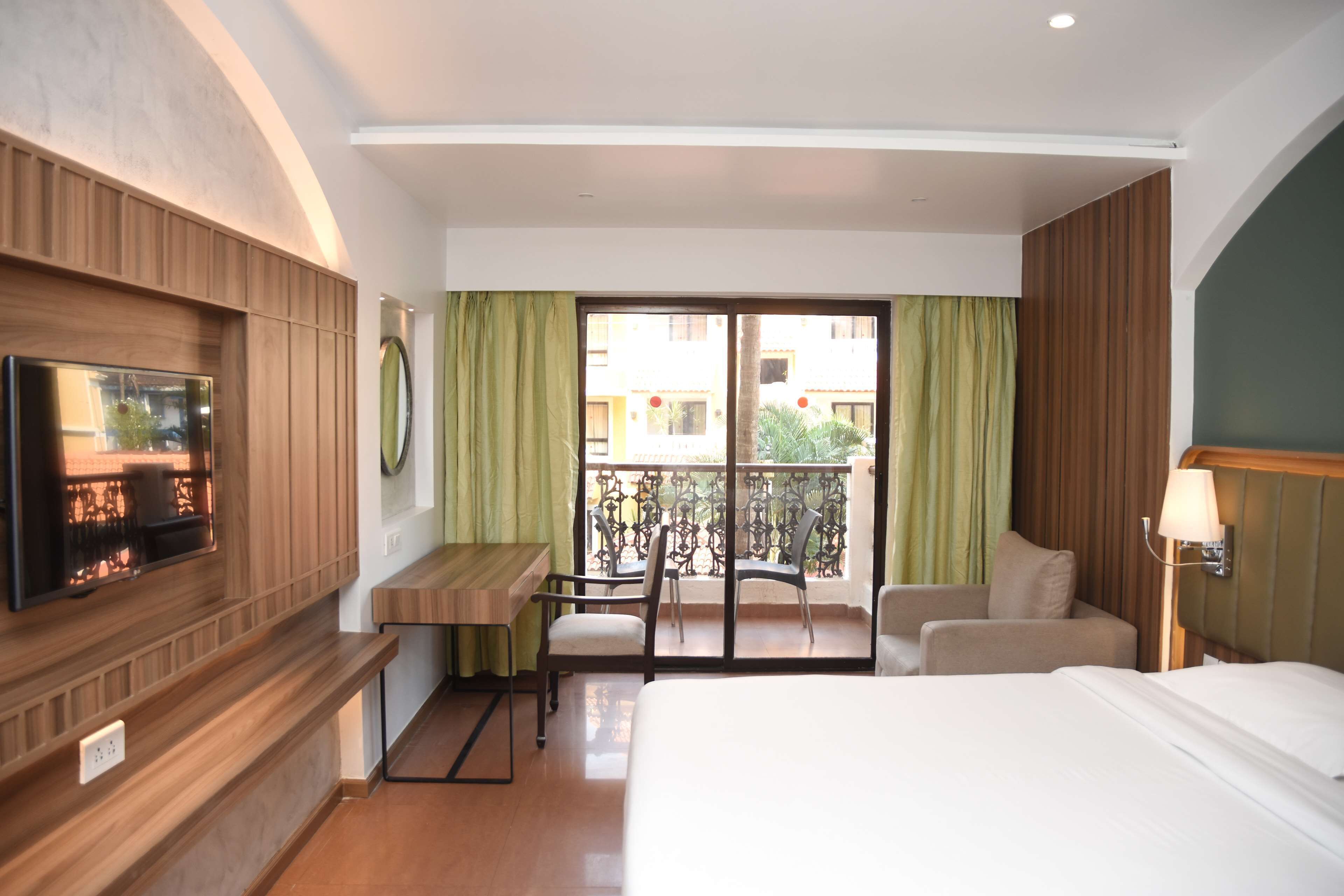 A hotel room with a bed, a desk, a chair, a TV, and a balcony with a view of buildings.